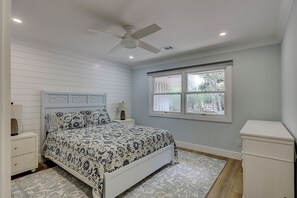 Une chambre à coucher comprenant un lit, une table de chevet, une lampe, un ventilateur de plafond, une fenêtre et une commode blanche.