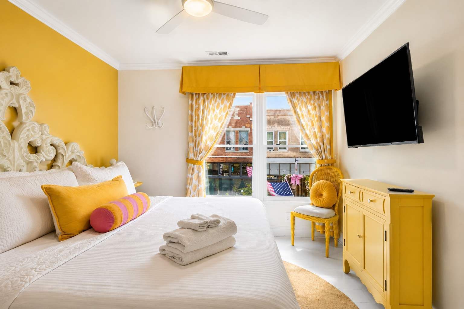 A hotel room with a bed, a television, a window with curtains, a yellow chair, and a yellow cabinet.