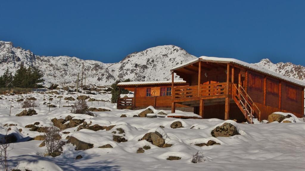 A wooden cabin with a balcony, surrounded by snow-covered ground and mountains.