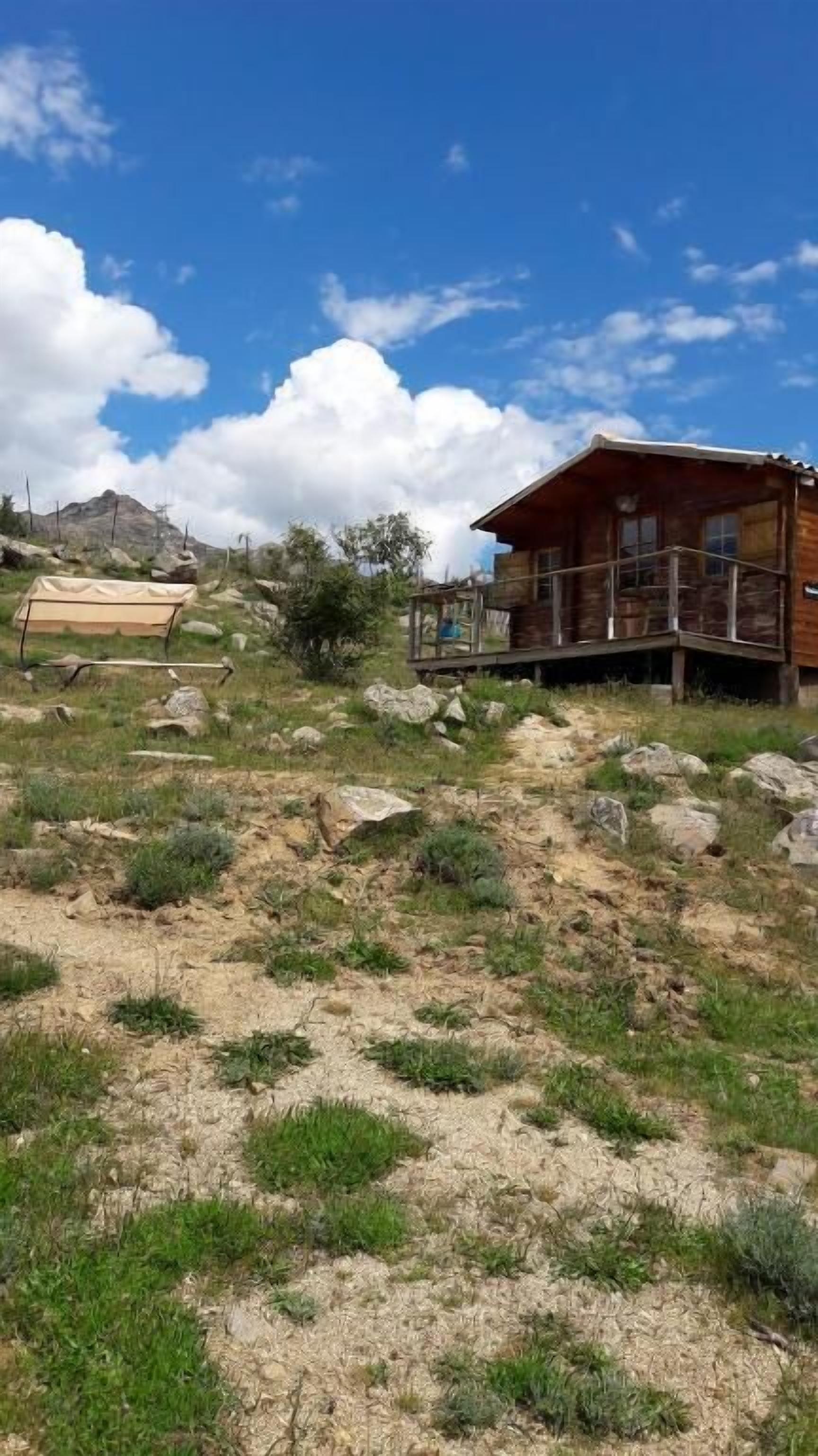 A wooden cabin with a balcony, surrounded by a grassy area with rocks and sparse vegetation.