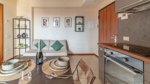 Een moderne keuken met een ingebouwde oven, een eethoek met een tafel voor twee personen, en een woonkamer met een bank en wanddecoratie.