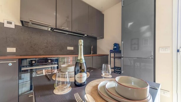Een moderne keuken met grijze kastjes, een inbouwoven en een eethoek gedekt met borden, kommen en een fles champagne.