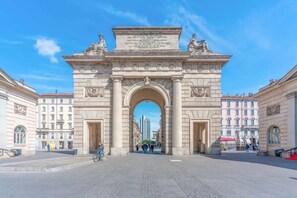 Ein großer, steinernes Bogenportal mit Inschriften, das von Gebäuden gesäumt wird und zu einem Platz führt.
