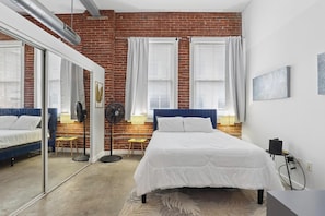 Un dormitorio con pared de ladrillo, cama con ropa de cama blanca, escritorio con silla y un espejo grande.
