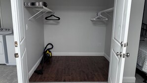 A closet with open doors, a vacuum cleaner, and a bed in the adjacent room.
