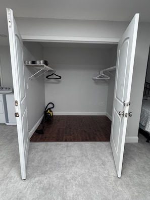A closet with open doors, a vacuum cleaner, and a bed in the adjacent room.