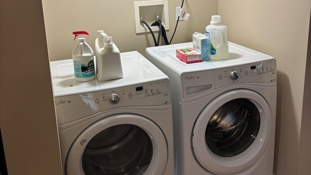 A laundry area with two front-loading washing machines, a white shelf with cleaning supplies, and a wall-mounted electrical outlet.