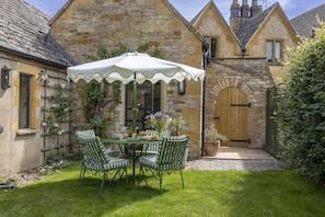 Ein Steinhaus mit einer Holztür, einer überdachten Terrasse mit Tisch und Stühlen sowie einer Rasenfläche.