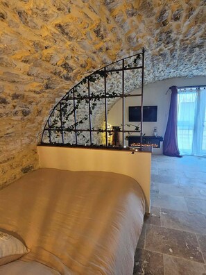 Ein Schlafzimmer mit Steinwand, einem Bett, einem Kamin und Blick durch ein Fenster.