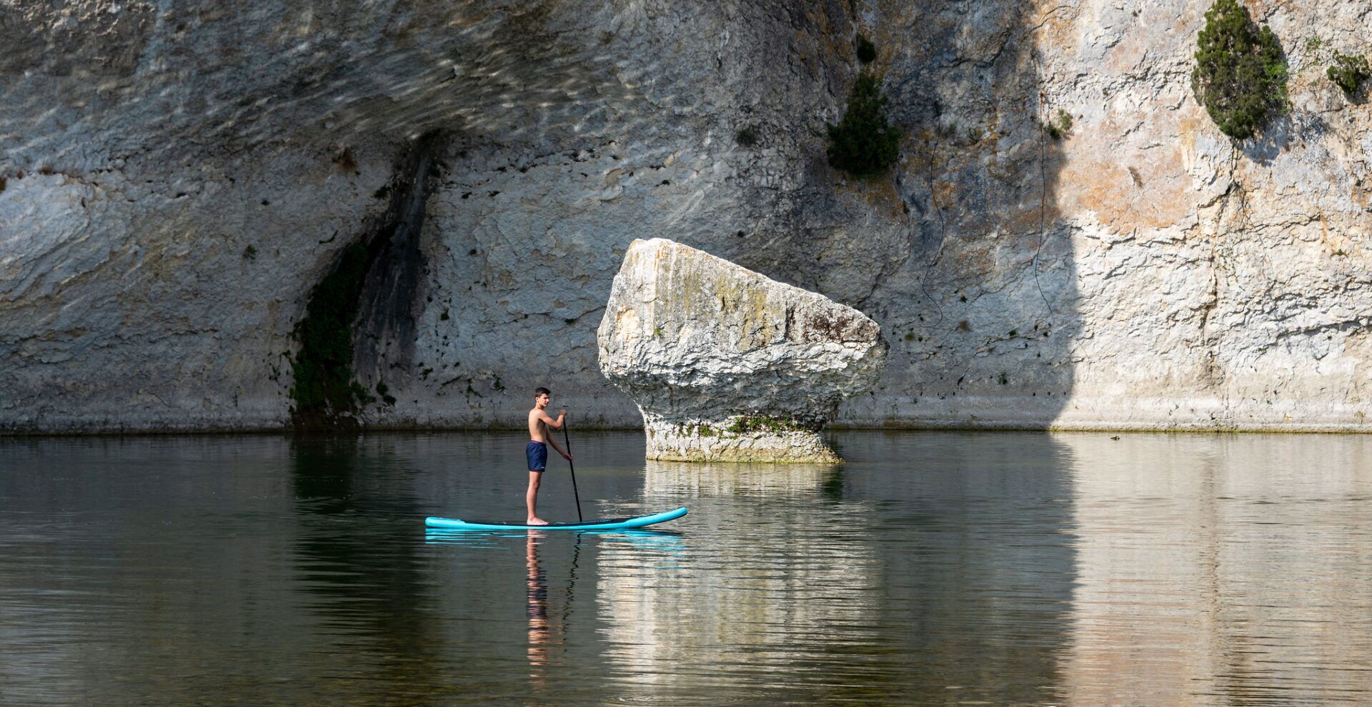 A person paddleboarding on calm water with a rocky cliff in the background.