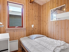 Ein Einzelbett mit weißer Matratze und Kissen, ein weißer Heizkörper, eine Holzwand und ein Fenster mit Jalousien.