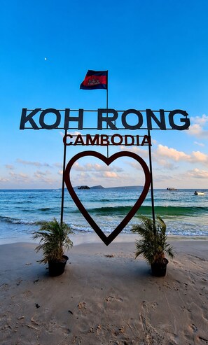 Escena de playa con un letrero con forma de corazón que dice 'KOHR RONG CAMBODIA' y una bandera.