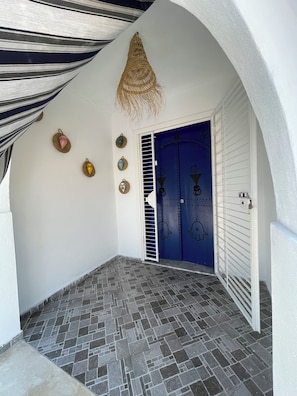 A covered entrance with a blue door, a tiled floor, and decorative wall hangings.