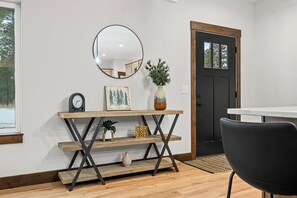 Un interior moderno con una consola de madera, un espejo redondo, un cuadro enmarcado, un florero con plantas y una silla negra.