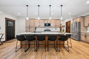 Una cocina moderna con una isla central, taburetes de bar, armarios de madera, electrodomésticos de acero inoxidable e iluminación colgante.