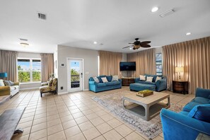 Ein geräumiges Wohnzimmer mit großem Fernseher, blauen Sofas, Deckenventilator und Blick durch die Glastür.