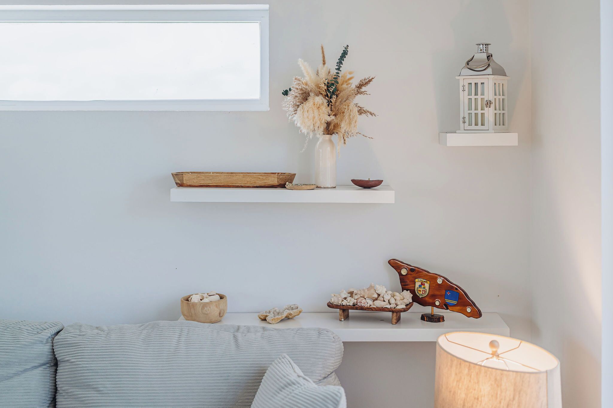 A minimalist living room with a white sofa, a wooden side table with decorative items, a white wall, and a window.