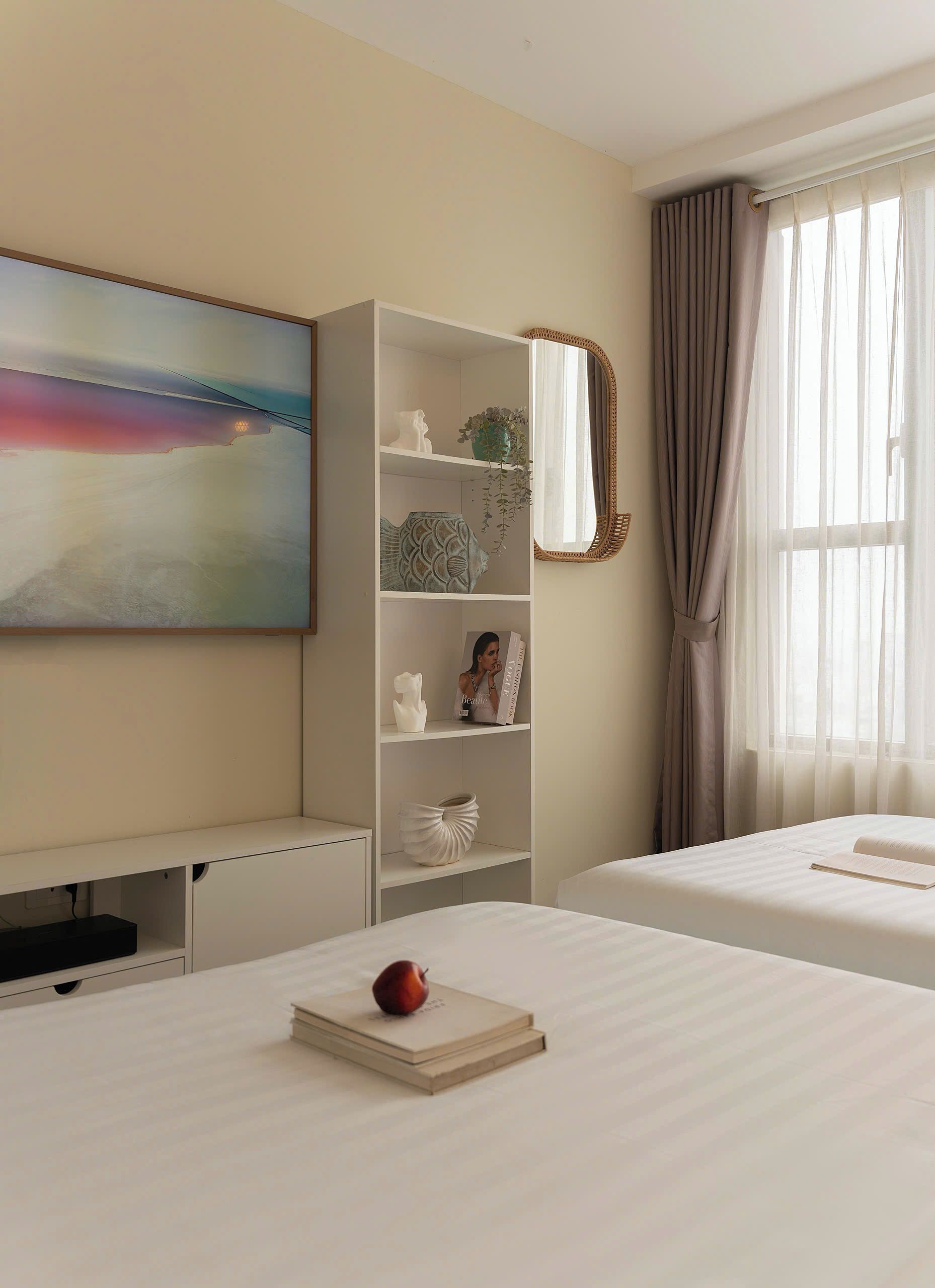 Une chambre d’hôtel avec un lit, une table de chevet sur laquelle se trouvent un livre et une pomme, une étagère blanche ornée d’objets décoratifs, un tableau accroché au mur et une fenêtre avec des rideaux.