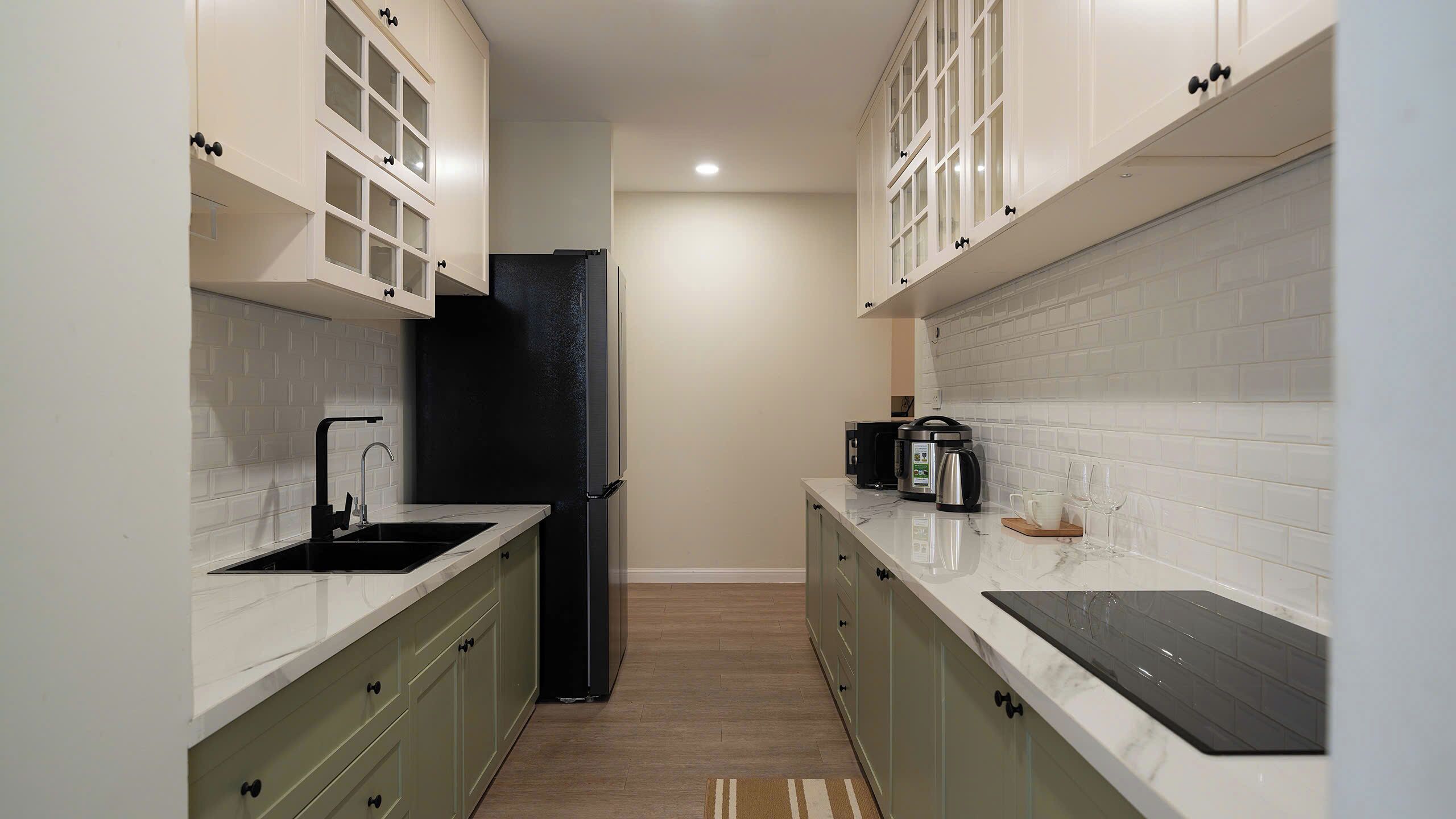 A narrow kitchen with white cabinets, a black refrigerator, a sink, and a countertop with a coffee maker and glassware.