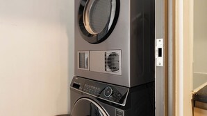 A stacked washer and dryer unit with a control panel, a wall-mounted power outlet, and a laundry basket. - Maison de Gu Tresor Residence (Ho Chi Minh City)