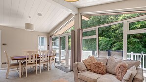 Un vaste espace de vie comprenant un canapé, une table à manger et des chaises, de grandes fenêtres offrant une vue sur la verdure, et un plafond avec des poutres apparentes.
