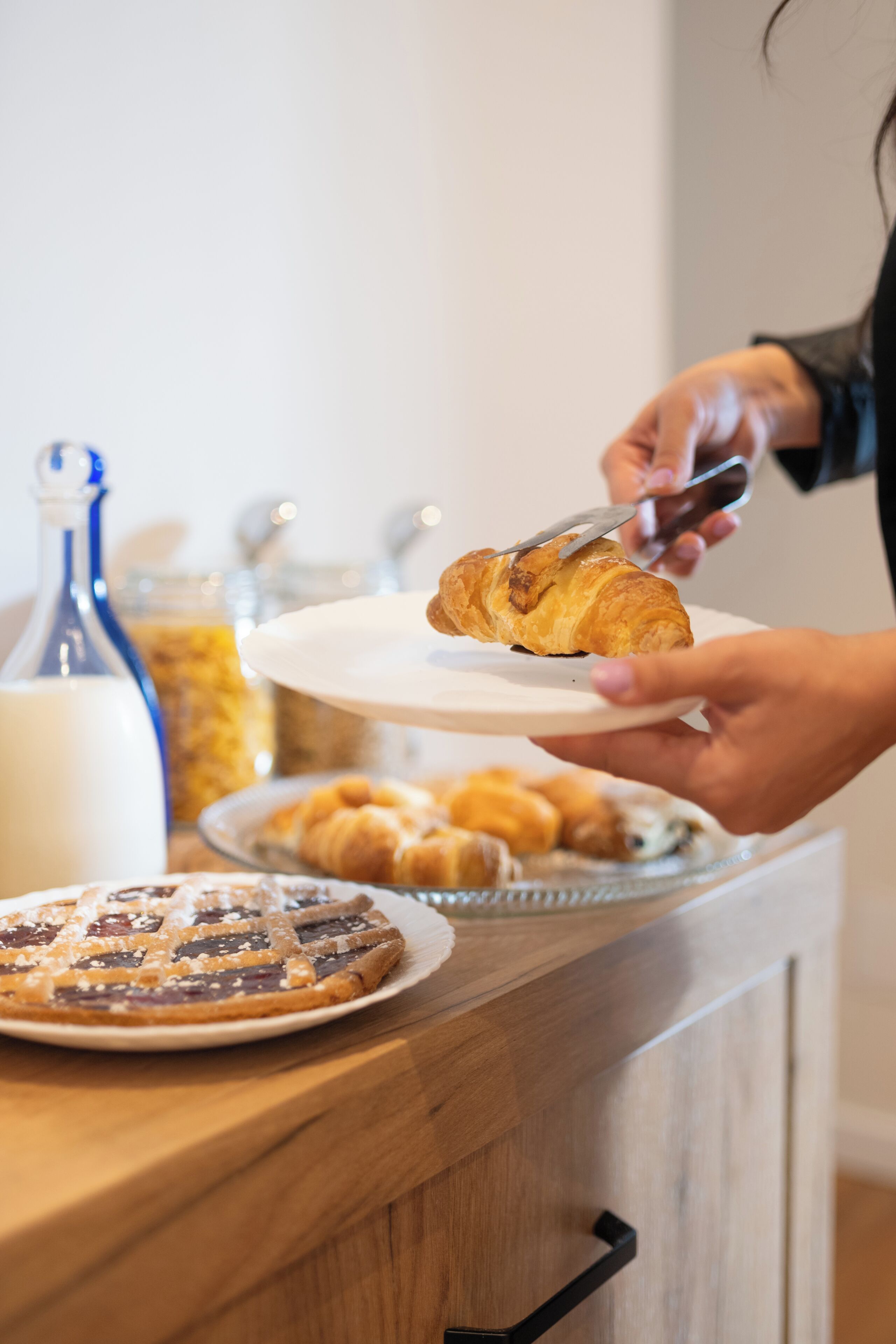A person holding a plate with a croissant, a jar of jam, and a bottle of milk on a wooden counter.