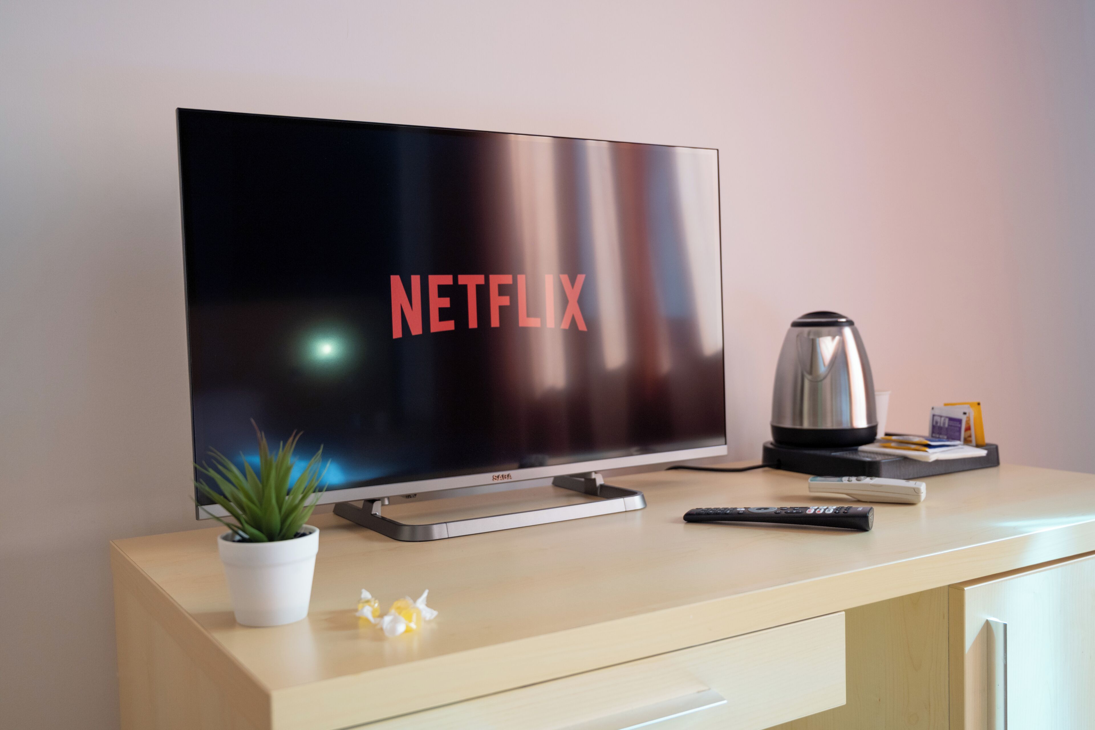 Un televisor de pantalla plana con el logotipo de Netflix, un control remoto, una cafetera y una planta pequeña sobre un escritorio de madera.