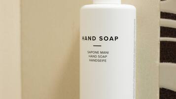 Uma garrafa branca de sabonete líquido com dosador, com a etiqueta 'HAND SOAP' e 'ORIENTAL TEA' sobre uma parede com azulejos bege.