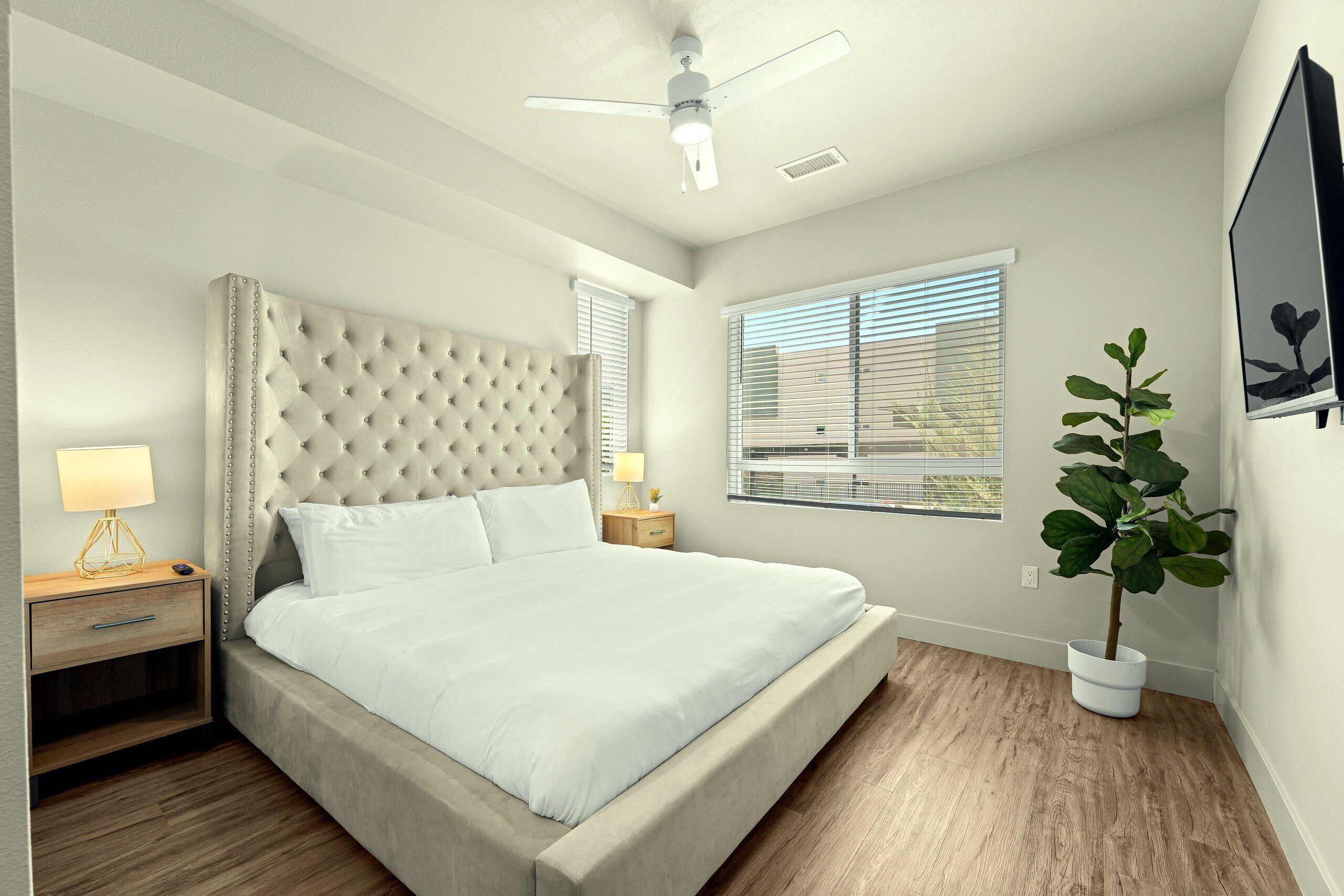 A bedroom with a tufted headboard, a single bed, a nightstand with a lamp, a potted plant, and a ceiling fan.