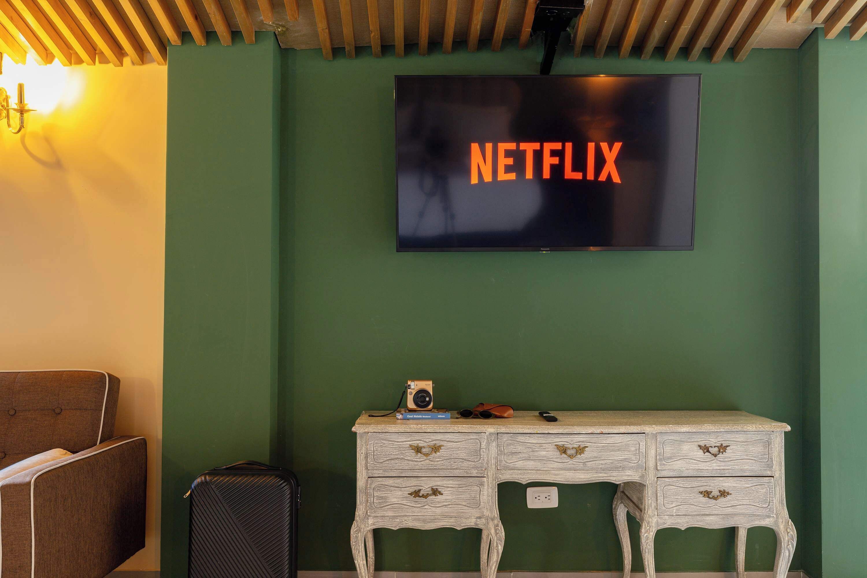 A room with a green wall, a white desk with drawers, a television displaying 'NETFLIX,' and a camera on the desk.
