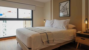 Een hotelkamer met een groot bed, een nachtkastje, een lamp, een ingelijst beeld op de muur en een raam met uitzicht op gebouwen.