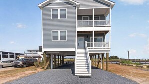 Une maison à deux étages avec un balcon à l’étage supérieur, un escalier menant à l’entrée et une vue sur l’eau en arrière-plan.