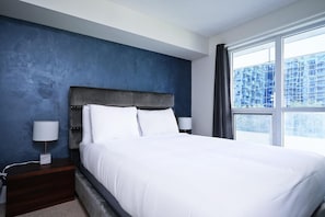 A bedroom with a large bed, a nightstand, a lamp, and a window with a view of buildings. - Northcrest Hotel (Toronto)