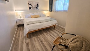 Un dormitorio con cama, mesitas de noche, una silla, un ventanal y un cuadro en la pared.