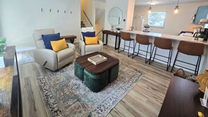 Una sala moderna con un sofá, reposapiés y una mesa de centro, un comedor con taburetes altos y una cocina con un amplio ventanal.