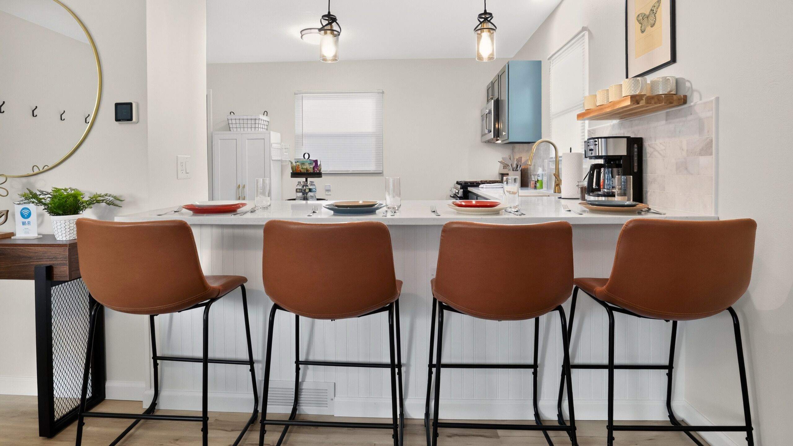 Uma cozinha moderna com bancada branca, quatro cadeiras marrons, uma máquina de café e uma janela com persianas.