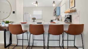 Una cocina moderna con encimera blanca, cuatro sillas marrones, una cafetera y una ventana con persianas.