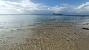 Ein Strand mit klarem Wasser, einer entfernten Insel und einem teilweise bewölkten Himmel.