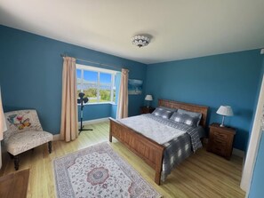 A bedroom with a bed, a chair, a nightstand, a lamp, a window with curtains, and a view of the outdoors.