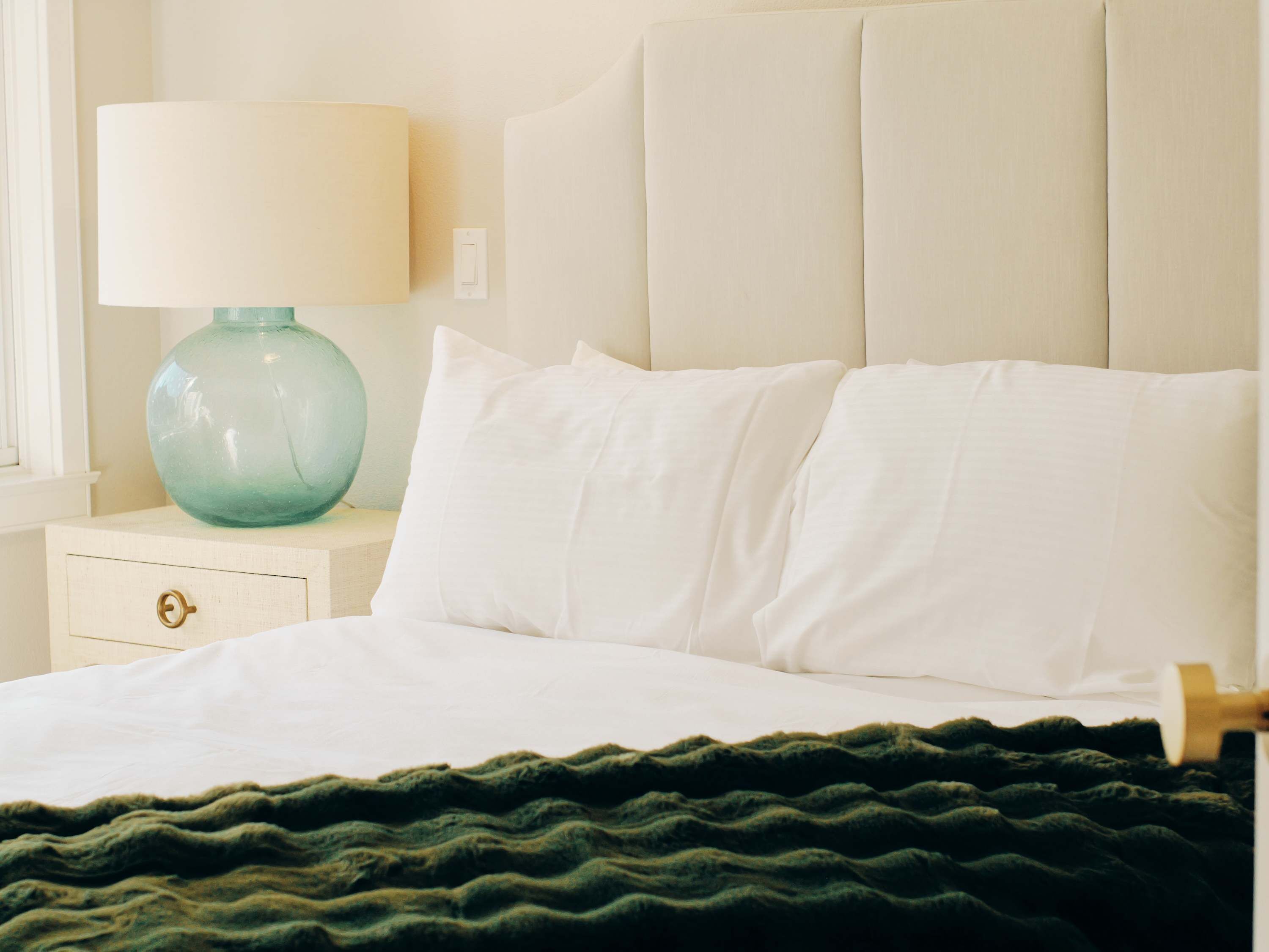 A bed with white bedding, a green textured blanket, a teal lamp, and a nightstand with a drawer.