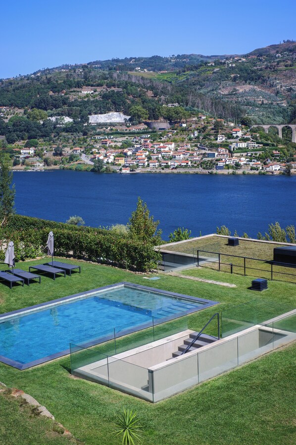 A swimming pool with a glass border, a grassy area with lounge chairs, and a view of a lake and hillside town.