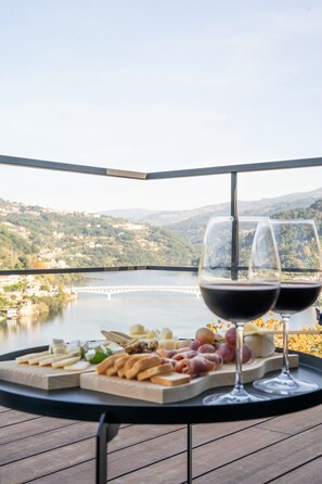 A table with a cheese board and wine glasses on a balcony overlooking a lake and hills.