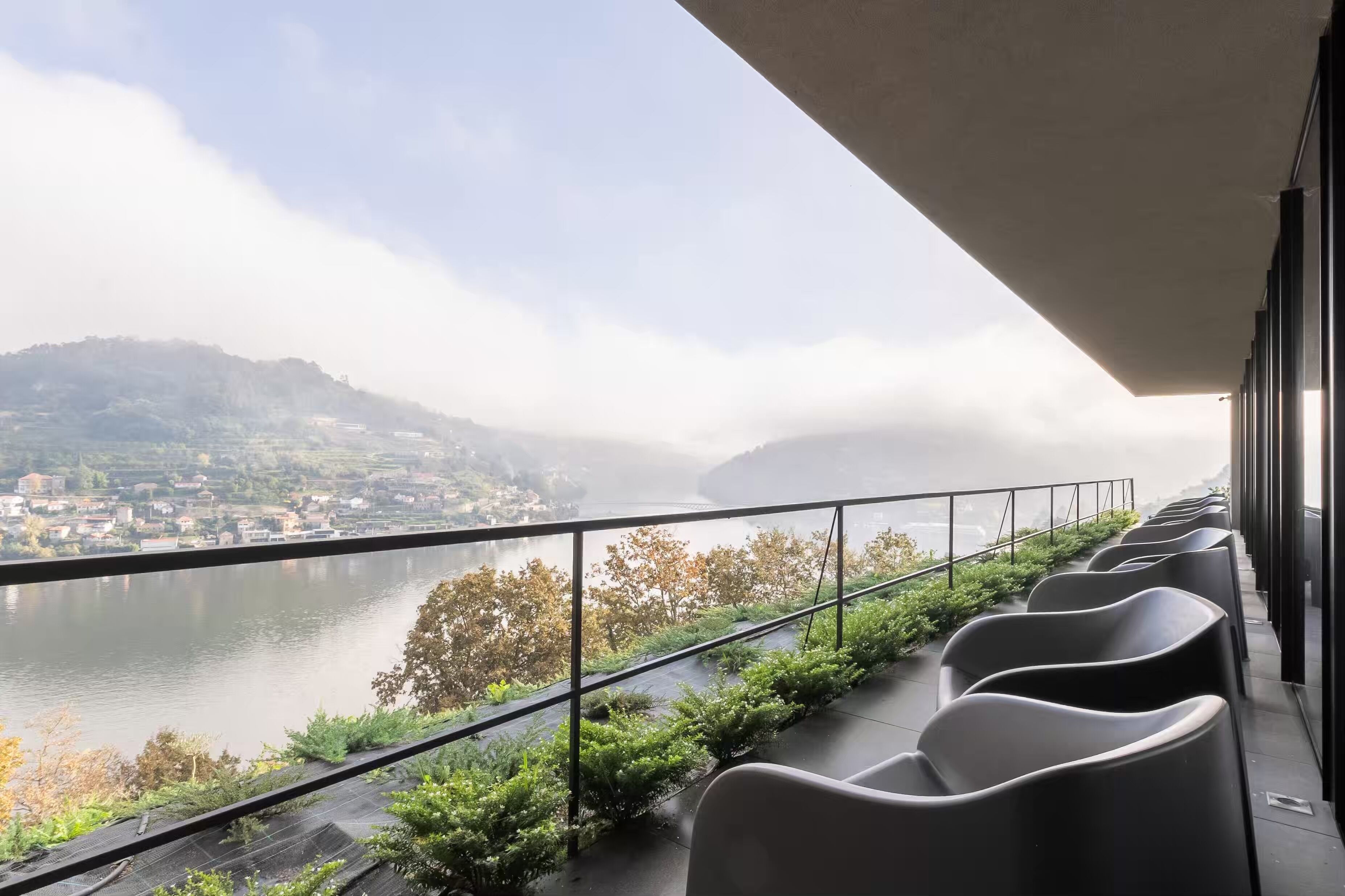 A balcony with a view of a river and a town, featuring modern chairs and a metal railing.