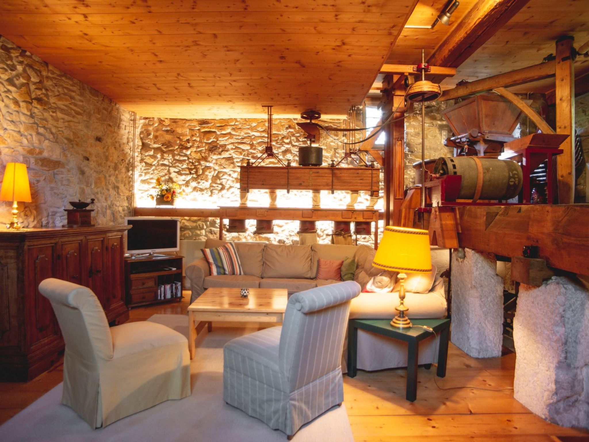 A cozy room with a stone wall, wooden ceiling beams, a sofa, armchairs, a television, and a fireplace.