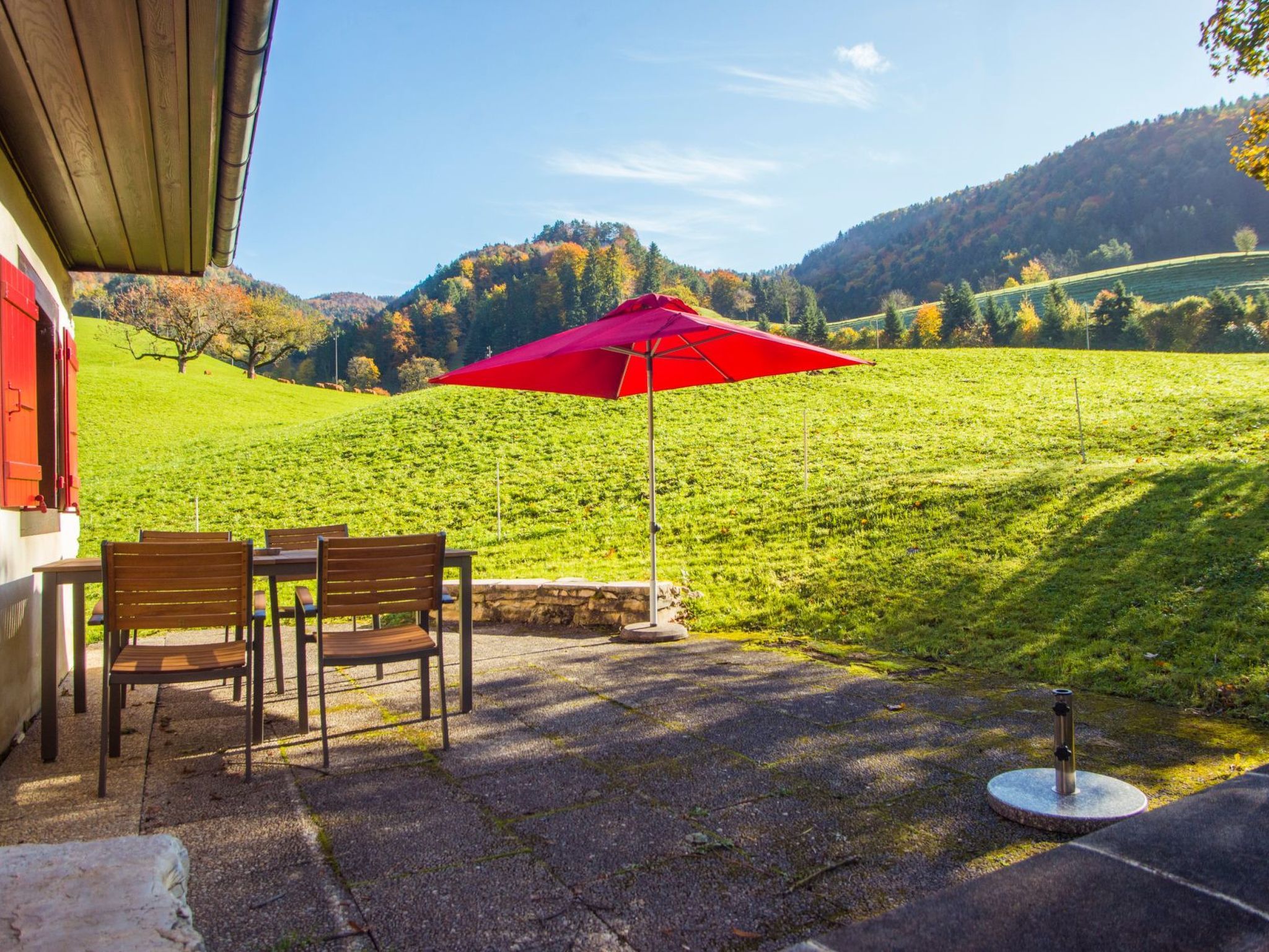An outdoor seating area with a red umbrella, a table, and chairs, overlooking a green field and hills.