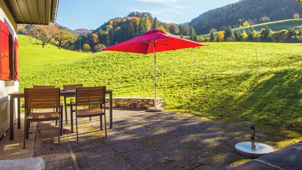 Ein Sitzbereich im Freien mit einem roten Sonnenschirm, einem Tisch und Stühlen mit Blick auf ein grünes Feld und Hügel.