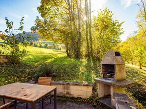 Un pati exterior amb un forn de pizza de pedra, una taula de fusta i cadires, envoltat de vegetació i arbres frondosos.