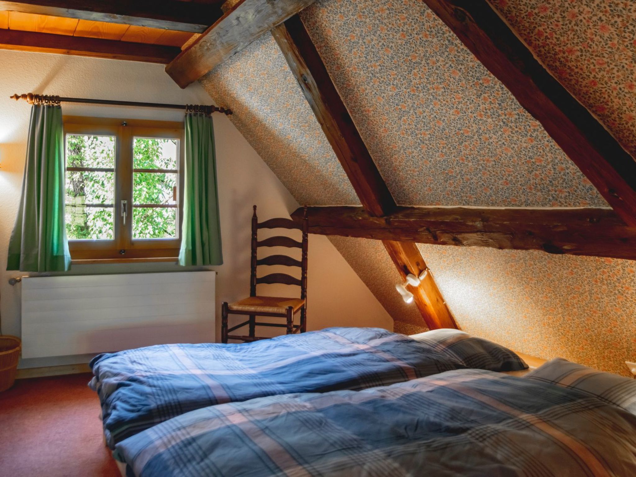 A bedroom with a sloped ceiling, a window with green curtains, a wooden chair, and a bed with a plaid blanket.