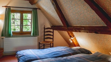 A bedroom with a sloped ceiling, a window with green curtains, a wooden chair, and a bed with a plaid blanket.