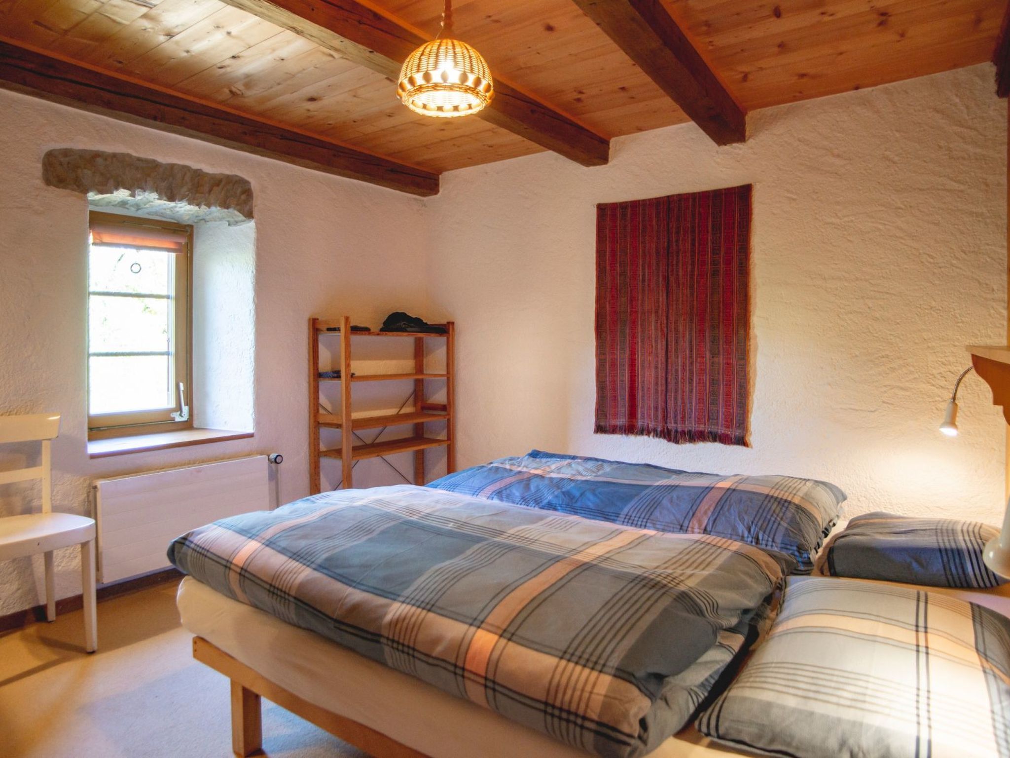 A bedroom with a wooden bed, a bench, a window, a radiator, and a hanging light fixture.